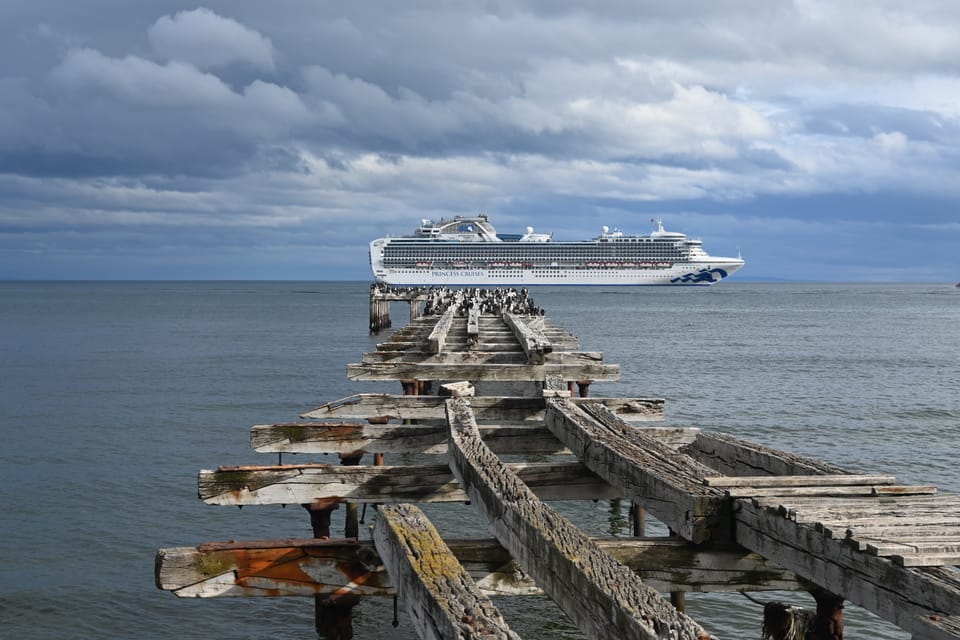 Alter Bootsanlegestege hinter dem ein Kreuzfahrtschiff vor Anker liegt. Am Ende des Steges sind Vögel zu sehen.